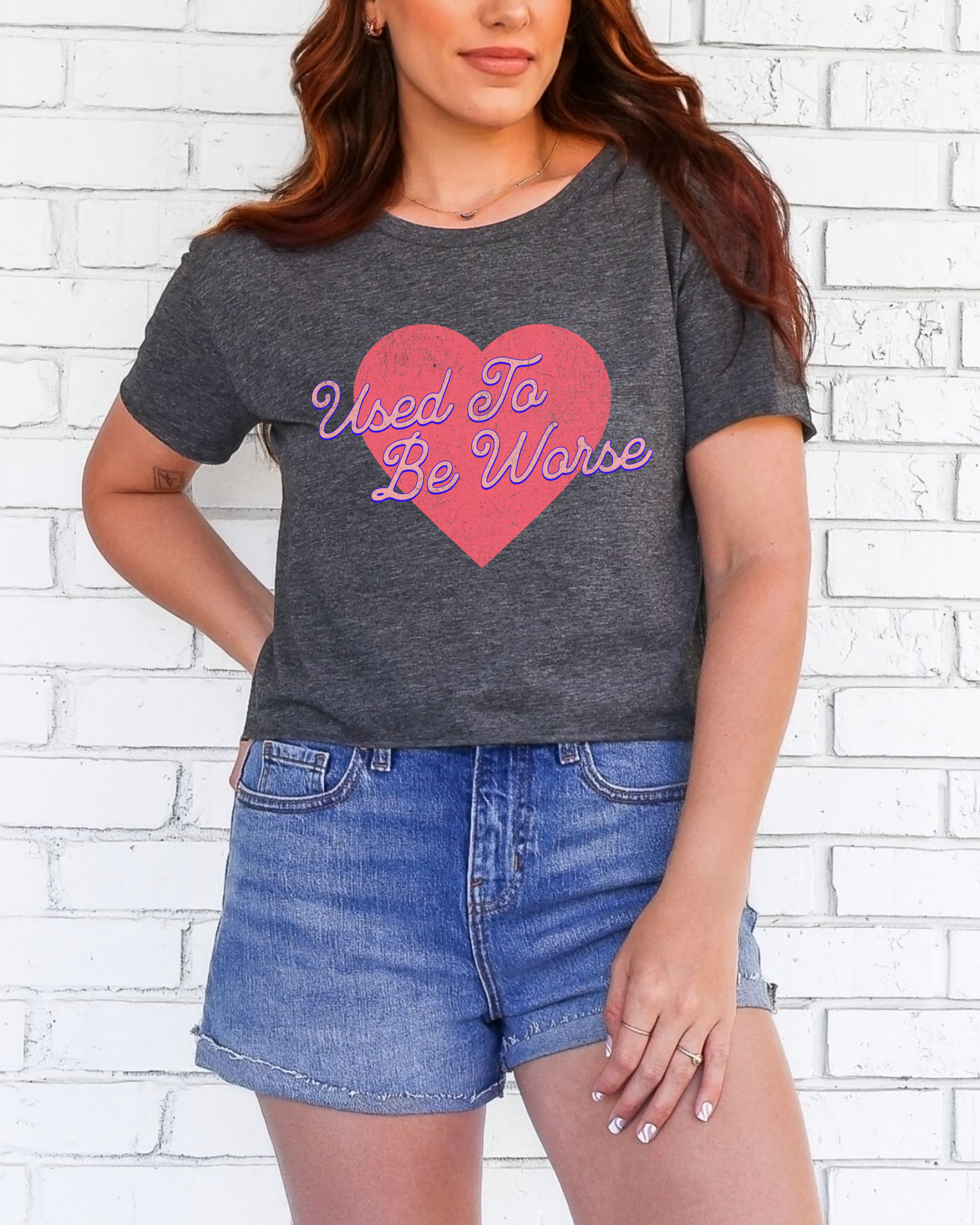 Woman wearing a dark gray t-shirt with a pink heart graphic and text, standing against a white brick wall.