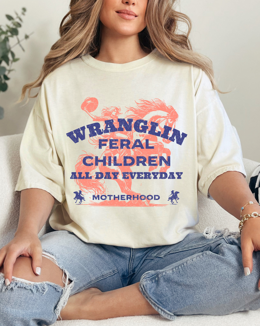 Person wearing a 'Wranglin' Feral Children All Day Everyday Motherhood' shirt sitting on a couch.