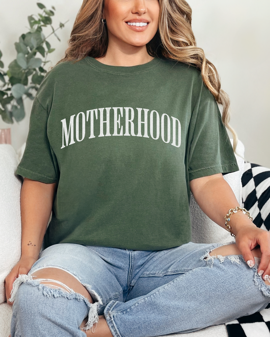 Woman wearing a green 'MOTHERHOOD' t-shirt sitting on a couch.
