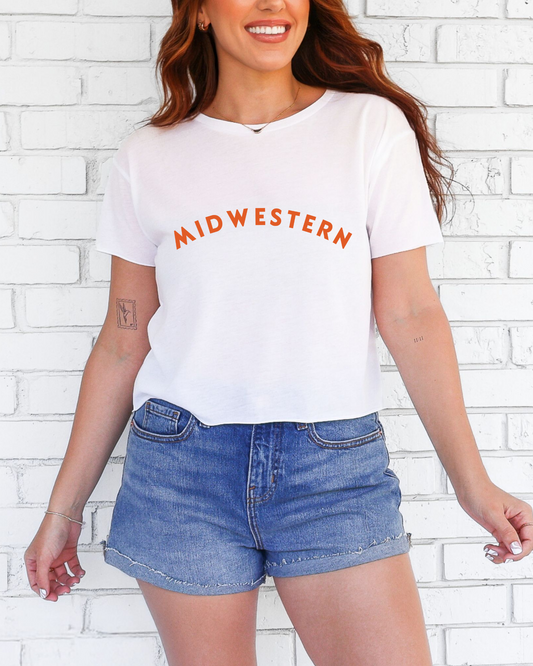 Woman wearing a white t-shirt with 'MIDWESTERN' text and denim shorts against a white brick wall.
